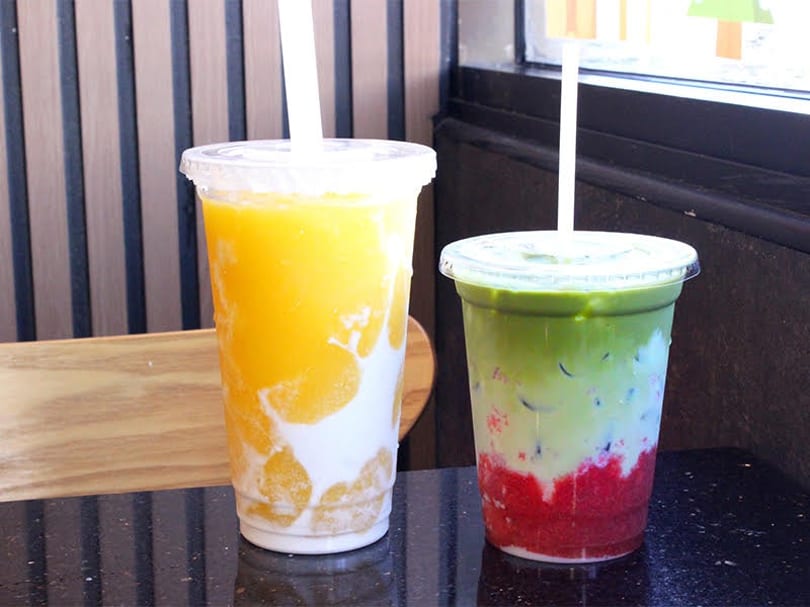 Two colorful layered drinks, one yellow and white, the other green, white, and red, on a dark counter.