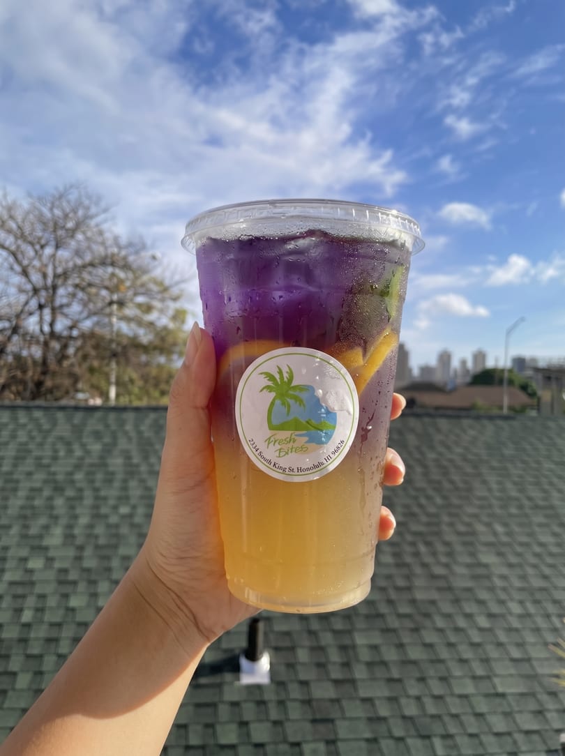 Hand holding a Fresh Bites drink with purple and yellow layers, against a blue sky.